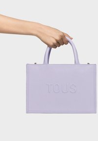 Borsa a mano in pelle lilla con logo "TOUS" impresso, design strutturato e doppi manici. Caratterizzata da una texture liscia e linee pulite.