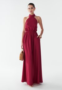 Vestido largo de cuello halter en rojo intenso, con cintura elástica, bolsillos y tela fluida. Los accesorios incluyen un bolso suave de color marrón.