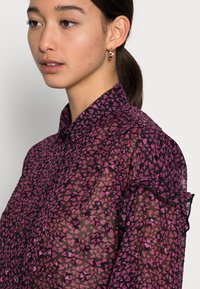 Camisa preta transparente com um padrão de corações cor-de-rosa, apresentando um colarinho, fecho de botão e mangas com babados.