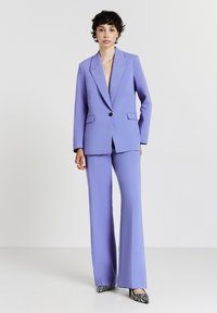 Donna in piedi con un tailleur lavanda composto da un blazer a un bottone e pantaloni a gamba larga, indossa scarpe con punta a stampa zebrate in bianco e nero.