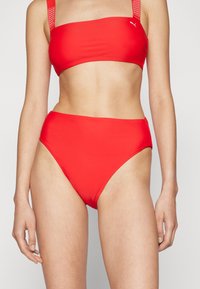 Rode twee-delige bikini gemaakt van een gladde stof. De top is een korte bandeau-stijl met brede bandjes, terwijl de onderkant een hooggesneden model heeft.