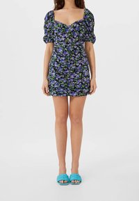 Blommig klänning med svart bakgrund och lila och gröna blommor, rynkad design, fyrkantig halsringning och puffärmar. Matchas med ljusblå sandaler.