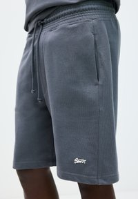 Pantaloni corti in cotone grigi con vita elastica, cordino, tasche laterali e un logo bianco sull'orlo inferiore. Tessuto liscio, design alla lunghezza del ginocchio.