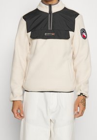 Krämmel fleecepullover med svart nylonpanel, kvartdragkedja i fronten och elastiska muddärm. Har logotypapplikation och ficka på framsidan.