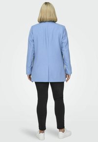 Blazer de color azul claro con un diseño entallado, con una abertura trasera sencilla, llevado sobre pantalones negros ajustados y zapatillas blancas. Textura de tela suave.