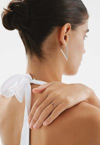 Pendiente geométrico de plata y anillo delicado de oro con acento azul, llevados por una persona con una blusa blanca sencilla con detalle de lazo. Textura de piel suave.