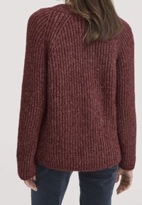 Femme portant un pull épais en maille côtelée bordeaux, vue de dos, associée à un pantalon bleu foncé, debout contre un fond uni.