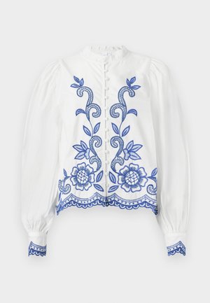 White blouse with blue floral embroidery, rounded collar, and puffed sleeves. Features a button closure and lace trim at the hem and cuffs.