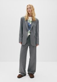 Blazer gri oversized cu rever în formă denotare și două butoane, asortat cu pantaloni gri largi. Tricou alb cu imprimeu sub blazer. Pantofi maro.