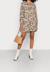 Beige Leopardenmusterkleid mit langen Ärmeln, einem V-Ausschnitt und rüschenförmigem Saum. Kombiniert mit einer hellbraunen Handtasche und weißen Stiefeln mit Absatz.