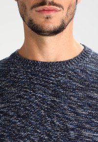 Pull en maille bleu marine avec un col rond et un motif texturé, présentant différentes nuances de fils bleus et blancs dans le tissu.