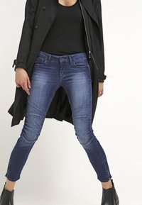 Blå denim skinny jeans med texturerade paneldetaljer, medelhög midja och blixtlåsdetaljer vid anklarna; bärs med en svart topp och en trenchcoat.