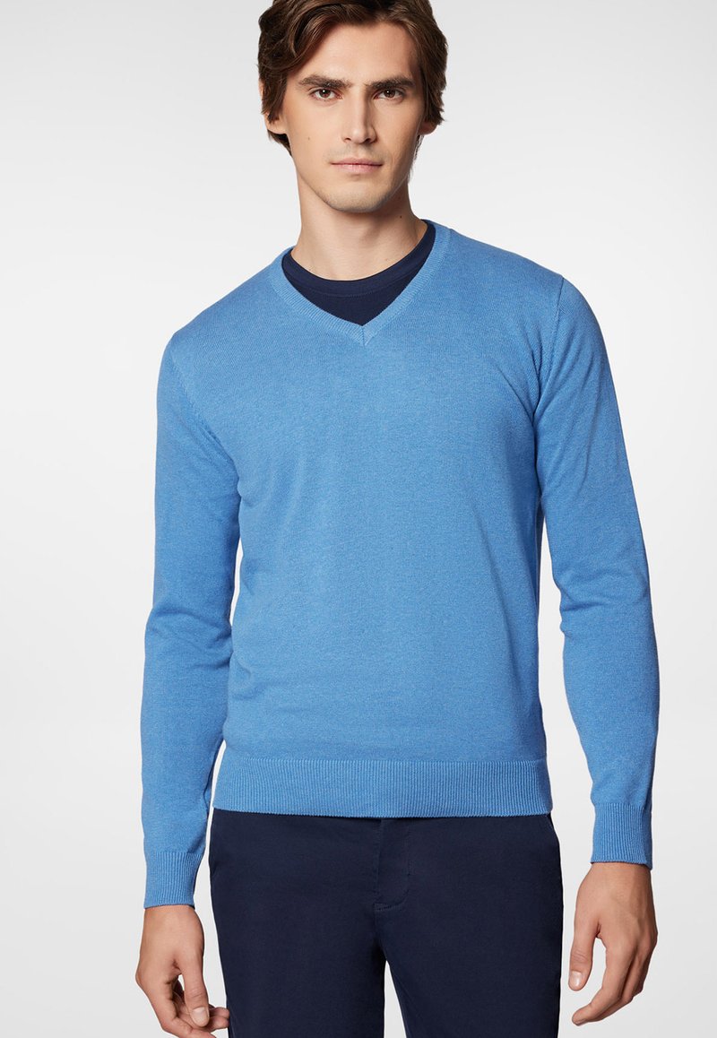 Niebieski sweter w serek wykonany z miękkiego dzianiny, z długimi rękawami oraz ściągaczami przy mankietach i u dołu, zaprezentowany na modelce na neutralnym tle.