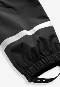 Pantalones impermeables negros con una banda reflectante plateada en la rodilla. Cuentan con cintura elástica y puños en los tobillos para un ajuste ajustable. Textura suave.