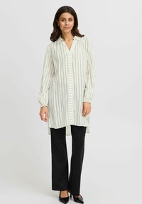 Blusa a righe chiare con colletto, chiusura con bottoni frontale e maniche lunghe, abbinata a pantaloni neri a gamba dritta e scarpe nere con punta.
