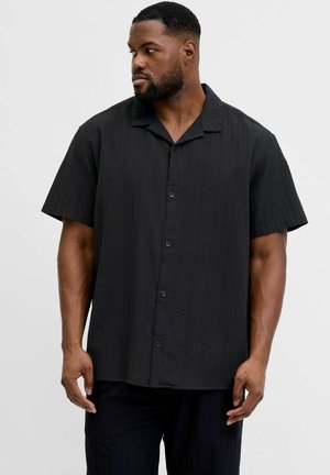 Bajszos férfi fekete, rövid ujjú, gombos ingben, balra nézve, egyszerű, világos színű háttér előtt áll.