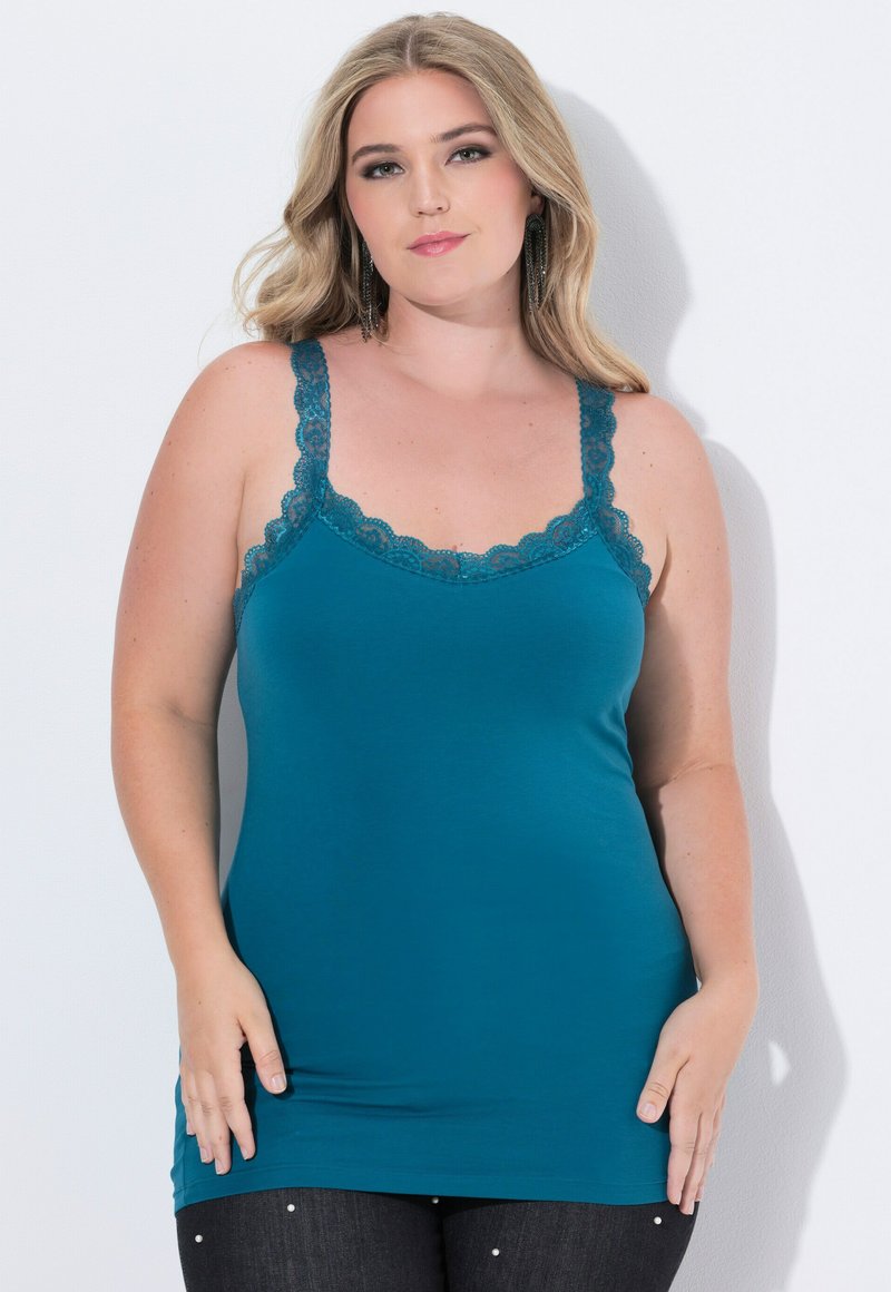 Donna con lunghi capelli biondi, che indossa un top senza maniche di pizzo turchese e pantaloni neri con piccoli chiodini bianchi, in piedi davanti a uno sfondo bianco.