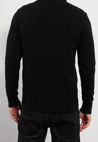 Pull noir en maille avec col rond, poignets et ourlet côtelés. Texture lisse ; coupe ajustée, vue de dos. Associé à un jean foncé.