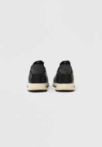 Paire de baskets décontractées noires et beiges vues de dos sur un fond gris clair uni.