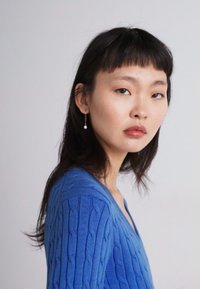 Suéter azul de punto trenzado con escote en V. El modelo tiene el cabello negro, lacio y largo, y lleva un pendiente de perlas colgante. Tela suave y texturizada.