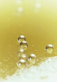 Klare vanndråper på en gulaktig væskeoverflate med fine, hvite partikler. Dråpene varierer i størrelse, reflekterer lys og skaper høydepunkter.