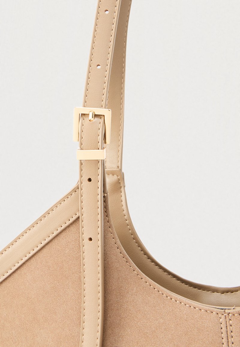 Bolso de mano de cuero beige con textura suave, correa ajustable y herrajes metálicos en tono dorado. Presenta un diseño elegante y costuras mínimas.
