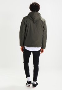 Olivengrøn hættejakke med en glat tekstur, lynlås lukning og sidelommer, parret med sorte slim-fit jeans og sorte sneakers.