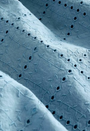 Jasnoniebieska tkanina z misternie wyszywanymi wzorami, z małymi wycięciami oraz ciemniejszymi akcentami. Tekstura wydaje się miękka i lekka.