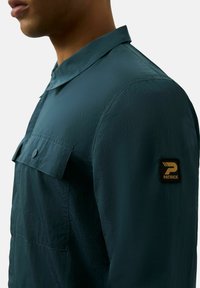 Ciemny zielony, długi rękaw, koszula z subtelną, pomarszczoną fakturą, kołnierzykiem, kieszenią na piersi oraz czarną łatą z złotym logo na rękawie.