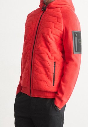 Persona che indossa una giacca a zip imbottita di un vivace rosso con cappuccio, tasche con zip nere e pantaloni neri, in piedi con le mani nelle tasche.