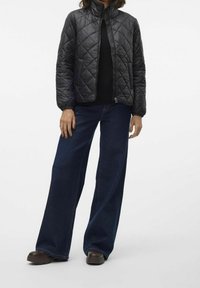 Černá prošívaná bunda s vysokým límcem, předním zipem a elastickými manžetami, doplněná širokými tmavě modrými džínami a hnědou obuví.