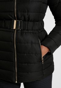 Černá péřová bunda s prošívanou strukturou, zlatými kovovými doplňky, nastavitelným pasem a kapsami na zip. Má přiléhavý střih.