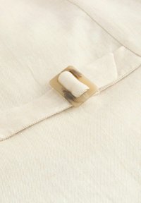 Ein hellbeiger Stoff mit einer horizontalen Rippstruktur, an dem ein quadratischer, heller Schildpattverschluss an einem Stoffgurt befestigt ist.