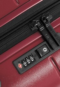 Bordeauxfarbener Hartschalenkoffer mit zwei schwarzen Reißverschlüssen, einem Zahlenschloss und einer strukturierten Oberfläche. Besitzt ein Logo am Reißverschluss-Zipper.