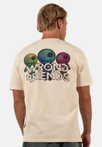 Beige t-paita, jossa on graafinen painatus kolmesta värikkäästä päästä ja teksti "VÄÄRÄT YSTÄVÄT" selässä. Puuvillamateriaali, pyöreä pääntie.