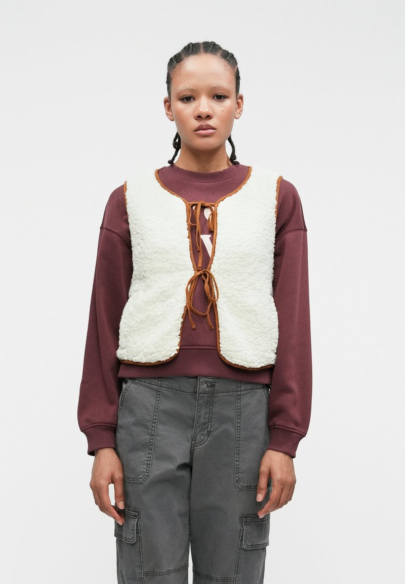 Fluffy witte vest met een bruine rand, met een sluiting aan de voorkant, gedragen over een wijnrood sweatshirt en gecombineerd met grijze cargobroeken.