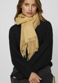 Écharpe en laine beige avec des bords frangés, drapée autour du cou d'une personne portant un pull noir. Texture douce, couleur chaude, design surdimensionné.