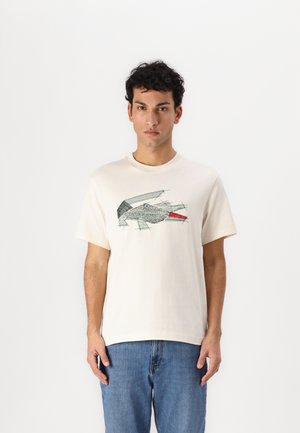 Camiseta de algodón color crema con mangas cortas, que presenta un diseño gráfico de un pez en verde y rojo. Corte casual, combinada con unos vaqueros azules.