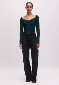 Langärmliges, tiefgrünes Oberteil mit Raffung, kombiniert mit schwarzen, hoch taillierten, weiten Jeans. Glattes Material, minimalistischer Stil, schwarze Schuhe.