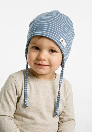 Kleinkind, das ein beigefarbenes Langarmhemd und einen blau gestreiften Hut mit Ohrklappen und Bindebändern trägt, lächelt sanft vor weißem Hintergrund.