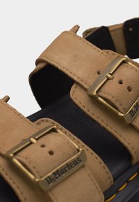 Κοντινό πλάνο από δερμάτινα σανδάλια σε χρώμα ταμπά με ρυθμιζόμενες μπρούτζινες αγκράφες και μαύρη επένδυση ποδιού με μαξιλαράκι, διακοσμημένη με κίτρινη ραφή.