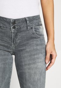 Šedé džíny z denimu mají dvojité zapínání na knoflíky, pečlivě zpracované švy a boční kapsy. Látka má mírně ošoupaný vzhled.