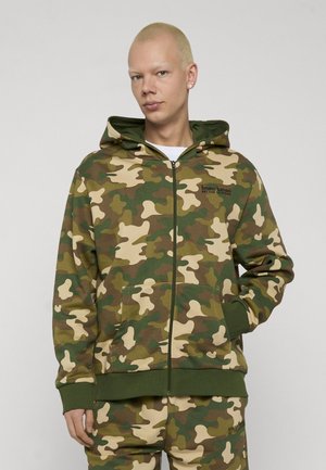Person trägt grüne und braune Camouflage-Kapuzenjacke mit Reißverschluss und passende Hose, steht vor einem schlichten hellen Hintergrund.