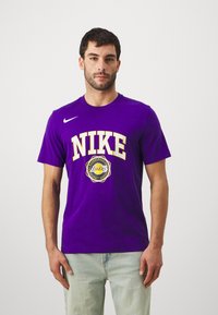 T-shirt en coton violet avec "NIKE" en jaune, un logo circulaire des Lakers et un col rond. Porté avec un jean en denim bleu clair.
