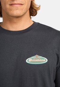 Camiseta de algodón gris oscuro con un colorido logo retro de Billabong en el pecho, con cuello redondo y cuello ribeteado.