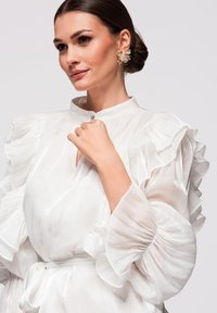 Femme aux cheveux foncés coiffés en chignon bas, portant un chemisier blanc transparent à volants et des boucles d'oreilles dorées à motifs floraux, tenant le col près du visage avec sa main.