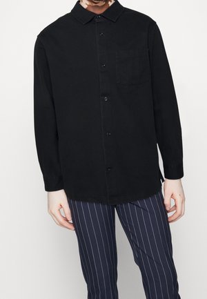 Chemise noire à manches longues avec un devant boutonné, col et poche poitrine, associée à un pantalon rayé bleu marine. Tissu lisse, sans motifs visibles.