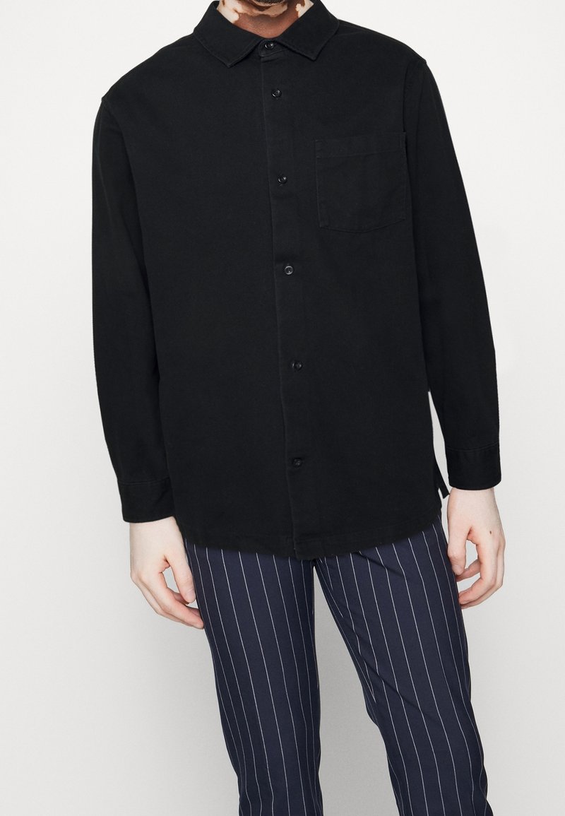 Chemise noire à manches longues avec un devant boutonné, col et poche poitrine, associée à un pantalon rayé bleu marine. Tissu lisse, sans motifs visibles.