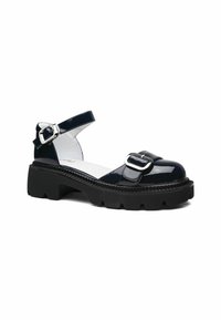 Sandalias de charol negro con una correa en el tobillo, suela negra gruesa y una correa frontal con un detalle de hebilla plateada.