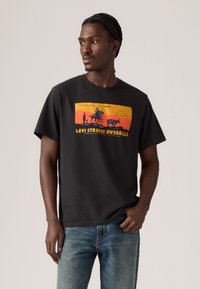 Mann med flettehår iført en svart Levi Strauss overall T-skjorte med et solnedgangs- og cowboygrafikk, kombinert med blå jeans.